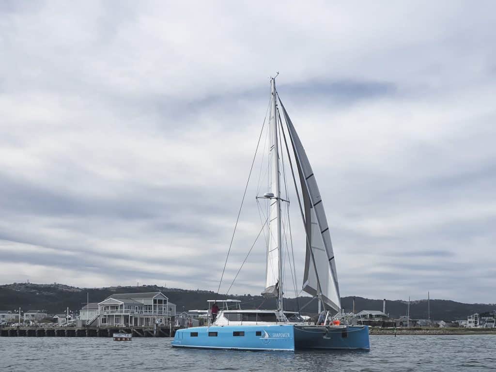 A bright blue Vision 444 catamaran named "Shapowza" sailing near a coastal town, with sails raised and a backdrop of houses and green hills under a cloudy sky.